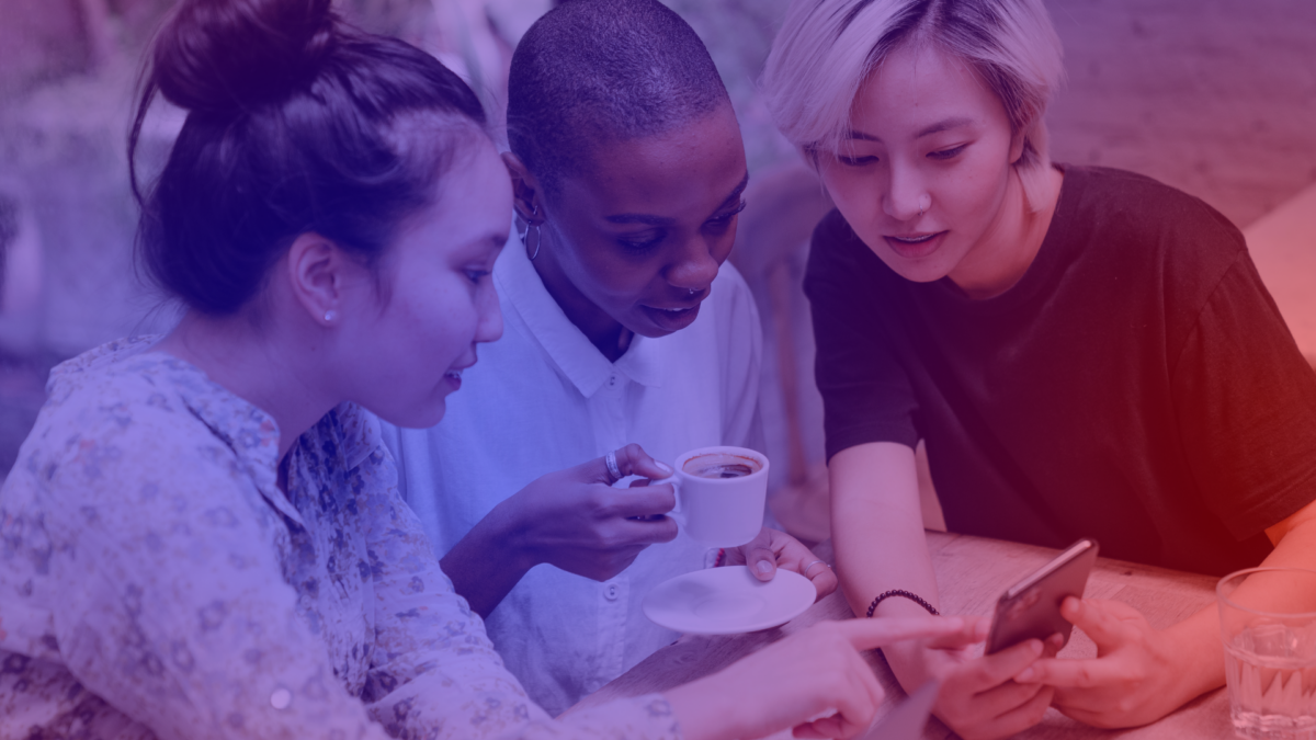 Drei junge Frauen betrachten gemeinsam ein Smartphone: Social Commerce in der Anwendung beim gemeinsamen Kaffee.