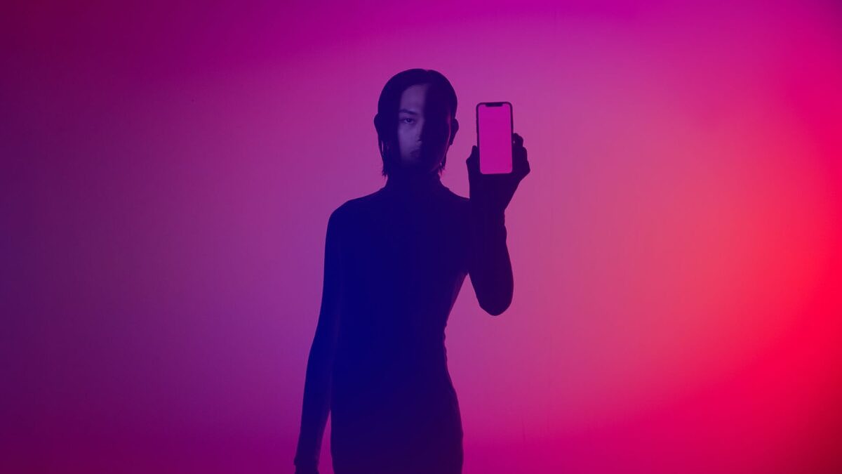 KI-Agenten: Person im Schatten hält ein Smartphone vor farbigem Verlaufshintergrund in Pink- und Violetttönen in die Kamera.